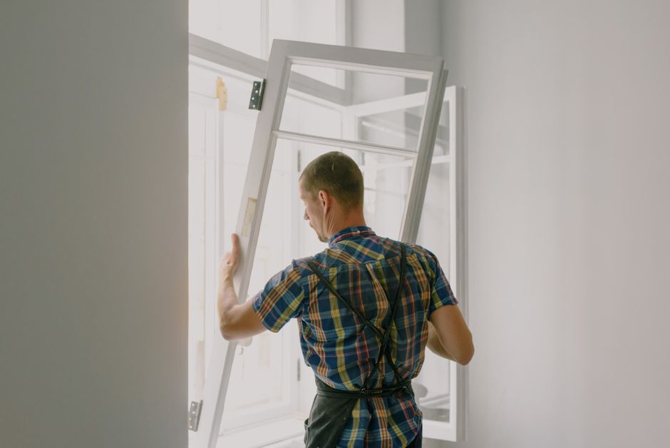 Back view of anonymous male fitter in checkered shirt and apron mounting window with hinges near white walls at home